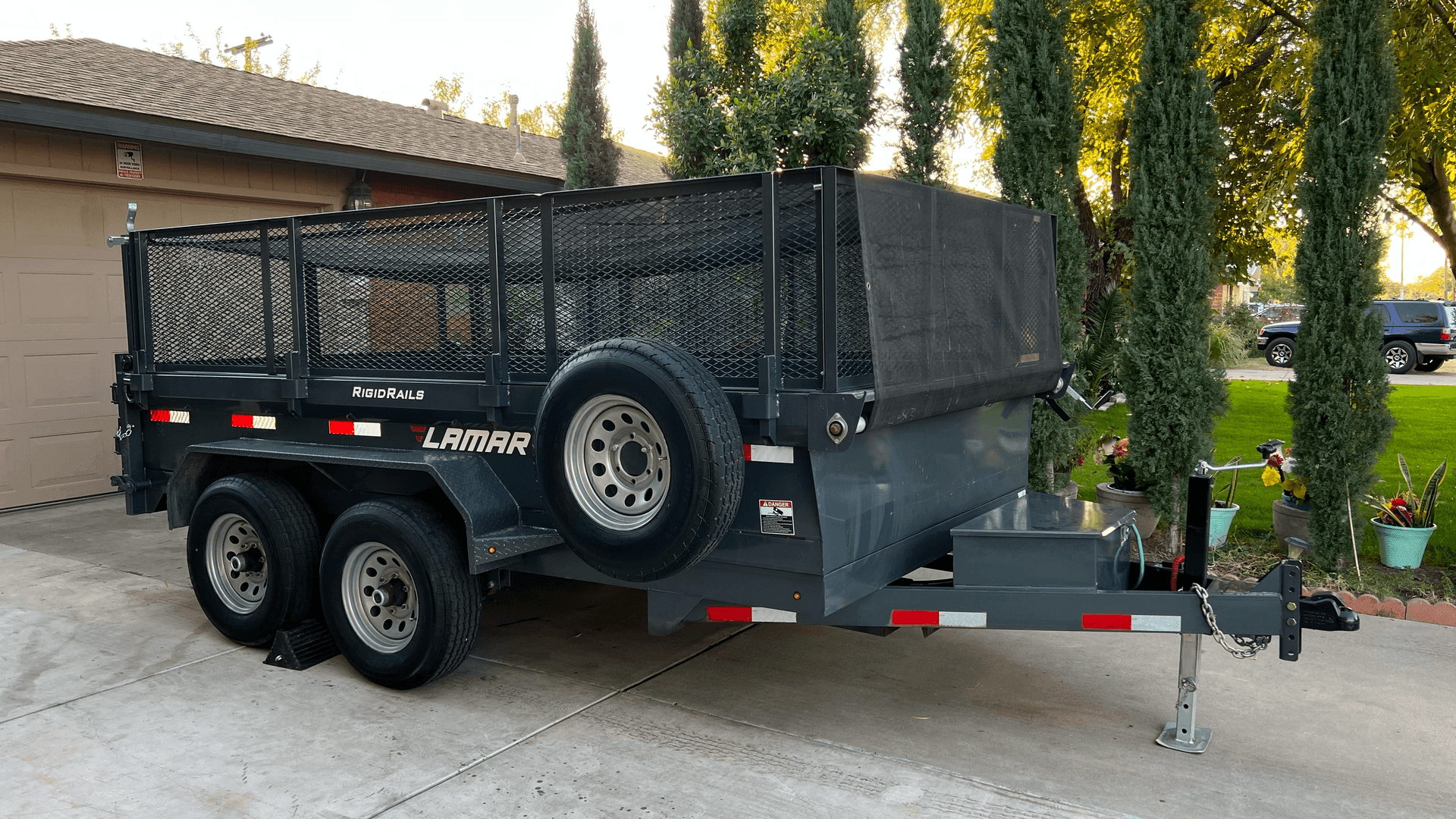A dark gray utility trailer parked on a driveway, surrounded by tall trees and greenery.